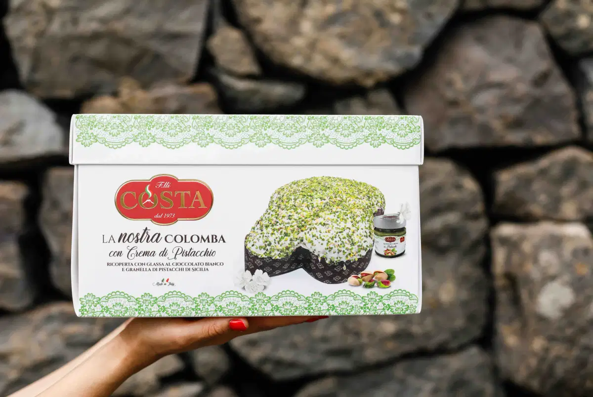 La confezione della colomba con crema di pistacchio tenuta in mano davanti a un muro di pietra lavica